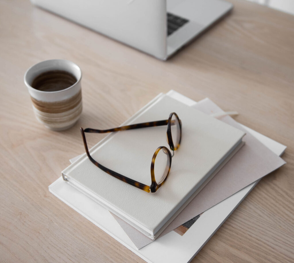 cup of coffee next to a stack of books with a pair of glasses on the top 
